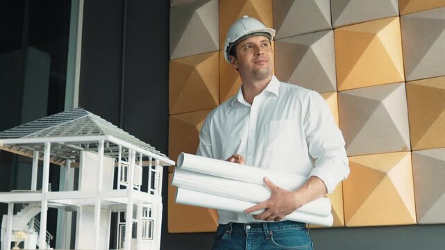 Professional caucasian project manager holding project plan with house model at table. Professional architect engineer wearing safety helmet while standing posing with confident. Manipulator.