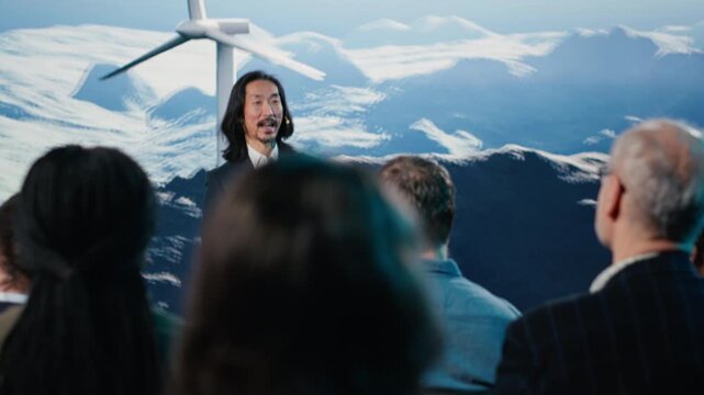 Asian man hosting renewable energy conference with windmill and turbine visuals. Businessman speaker delivers pitch to mixed age audience, highlighting sustainable wind power in auditorium. Camera B.
