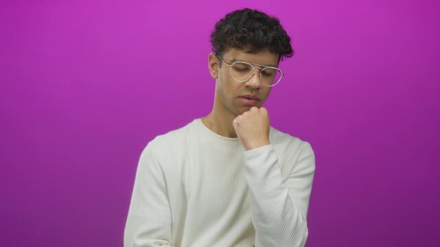 Young hispanic man wearing glasses holds his chin with his hand in studio scene; pensive reflection.