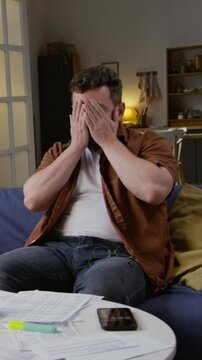 Vertical shot of exhausted adult man closing computer while sitting on sofa and working from home various documents scattered on table