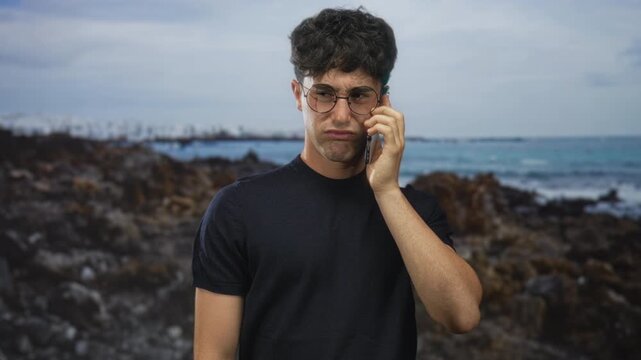 Man holding phone to ear on rocky seaside beach wearing glasses with bare forearm visible and dark t shirt; concern uncertainty.