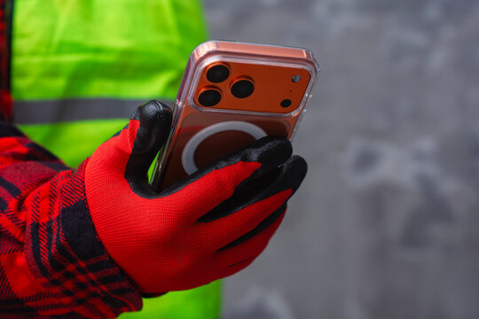 Worker hands wearing protective gloves holding smartphone at construction site. Concept of mobile technology in construction industry, digital tools, communication and modern industrial work.