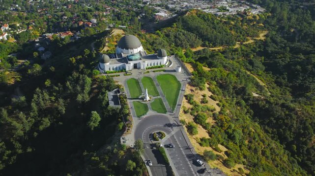 Aerial drone view of Griffith Observatory on Mount Hollywood with green lawns, surrounding hills and expansive Los Angeles California cityscape under clear daylight sky.