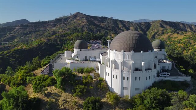 Cinematic aerial view of Griffith Observatory surrounded by green hills. White landmark rises above Los Angeles, showing contrast between architecture nature and vast California landscape.