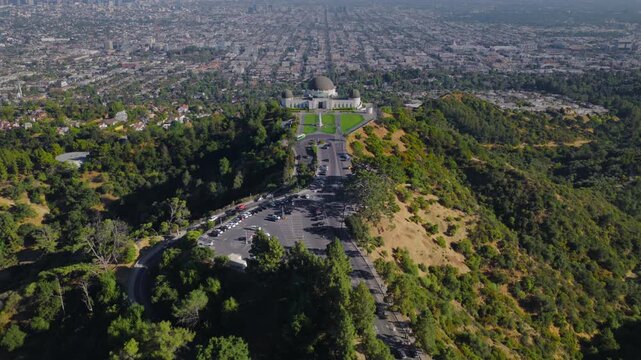Drone aerial view of Griffith Observatory from the front looking toward dense Los Angeles California city grid with green park lawns, roads, cars and clear sky.