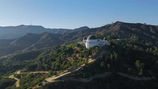 Drone aerial side view of Griffith Observatory surrounded by rugged Hollywood Hills with winding trails, green slopes and bright California daylight.