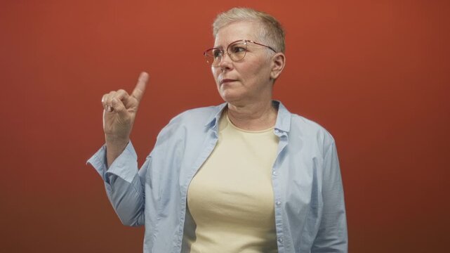 Woman pointing finger in orange studio wearing glasses and casual shirt; confidence assertiveness decision.