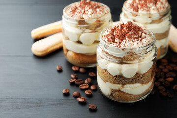 Delicious tiramisu in jars, savoiardi biscuit cookies and coffee beans on dark wooden table, closeup. Space for text