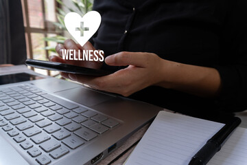 A businessman holding a cell phone with a heart icon with a cross and the word "Wellness." He holds a laptop in front of him. Health app concept.	