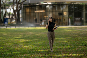 Fototapeta premium Businesswoman walking in park multitasking with smartphone and clipboard