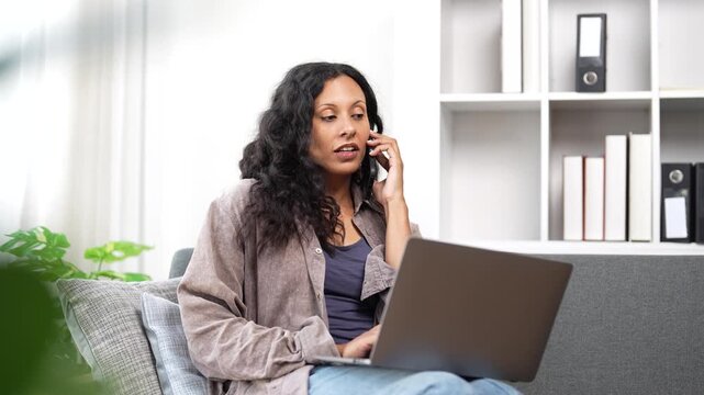 Cheerful female entrepreneur sitting on the sofa with a laptop, managing her business and talking on her smartphone with a customer, enjoying the flexibility of remote work in her living room