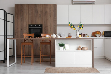 Fototapeta premium Interior of kitchen with counter, chairs, oven, teapot and artificial lemons branches in vase on table