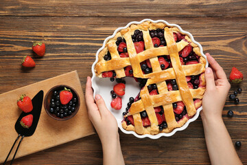 Female hands and baking dish with tasty berry pie on wooden background