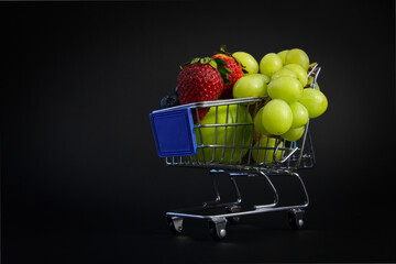 Miniature shopping cart filled with fresh organic fruits on black background
