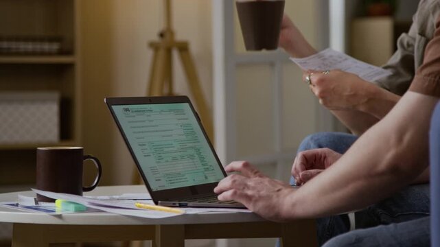 Side view close up shot of unrecognizable couple filling in online tax form via laptop with focus on hands holding documents, copy space