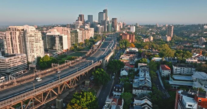 Australia, Sydney urban landscape with large truss bridge carrying road and rail traffic, a multi-lane highway, suburban streets, and towering city skyline in distance under clear sky. Aerial flight