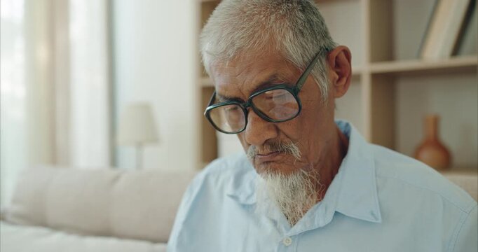 Elderly Asian man with white goatee on sofa in sunlit living room, senior retirement lifestyle and mindful healthy aging in natural light	