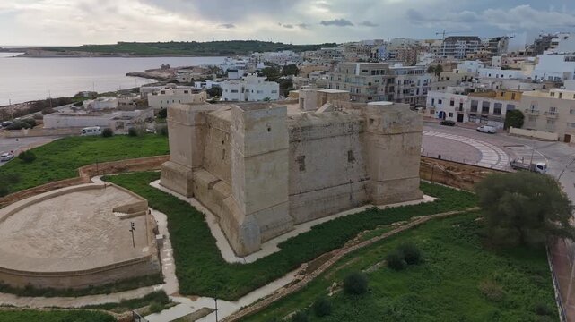 Orbiting drone view of Torri San Tumas, a 17th-century Wignacourt tower in Marsaskala, Malta, showcasing its historical limestone architecture amidst coastal and urban Mediterranean scenery.
