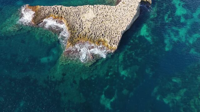 Low-level drone flyover above It-Taqtiegħa island near Triq Xrobb L-Għaġin, showing limestone rock formations surrounded by crystal-clear turquoise Mediterranean waters along Malta&rsquo;s southern coast.