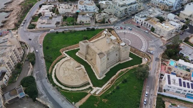 Orbiting aerial view of Torri San Tumas, a historic watchtower in Marsaskala, Malta, surrounded by urban landscapes, showcasing its medieval architecture and cultural significance.