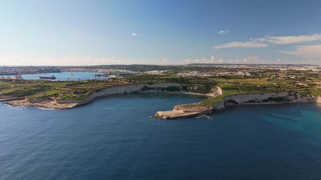 Aerial view of southern Malta, capturing rugged cliffs and a calm bay on the Mediterranean waters, while the Birżebbuġa and Marsaxlokk cityscape and harbor stretch across the horizon on a sunny day.