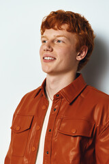 Obraz premium Young man with red hair wearing brown leather jacket smiling and looking aside with white background for portrait and casual fashion style.