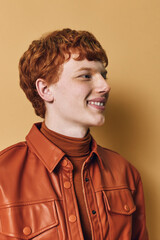 Obraz premium Young man with red hair and freckles smiling in brown leather jacket and turtleneck sweater against beige background in studio portrait.