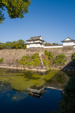 A traditional white castle turret stands atop ancient stone ramparts overlooking a calm moat in Osaka, Japan. Bright sunlight and clear blue sky create vivid reflections on the water, with lush