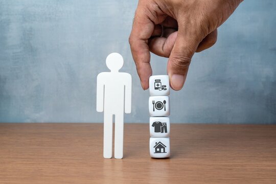 Hand arranging blocks with basic human needs icons symbolizing shelter, clothes, food, and medicine beside a human figure. Essential life requirements, human rights, and social welfare concept.