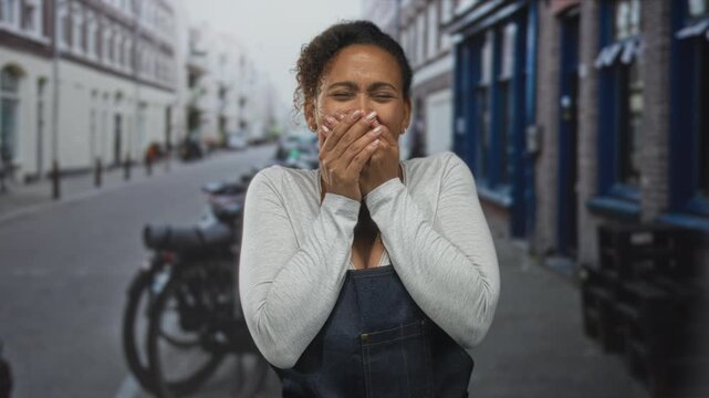 Woman covering mouth with hands on a cobbled street with parked motorcycle and row of buildings; surprise joy.