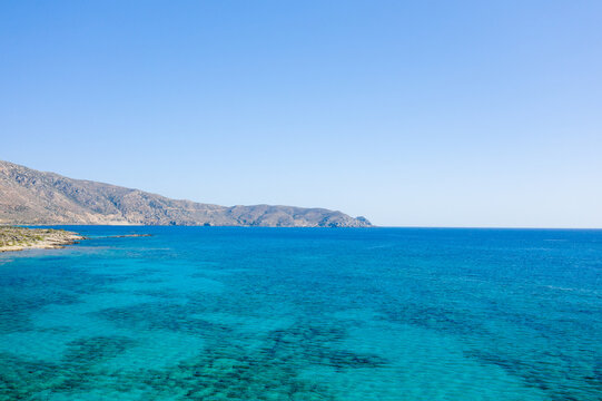 Expansive view of clear blue Mediterranean waters meeting a rocky shoreline and distant arid hills under a wide, cloudless sky. The tranquil seascape captures the vivid colors and open space of the