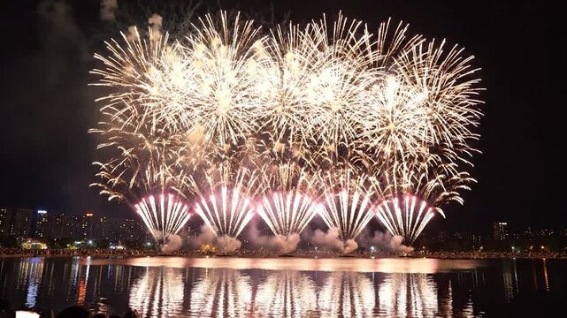 Dazzling fireworks erupt over a reflective body of water at night with a city skyline backdrop