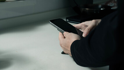 Close-up of Hands Using a Smartphone in a Quiet Kitchen with Soft Morning Light （柔らかな朝の光が差し込む静かなキッチンで、スマートフォンを両手で操作する様子）