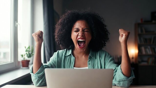 An overjoyed African American woman celebrates a huge success while sitting in front of her laptop at home.