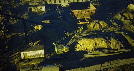 Fototapeta premium Heavy machinery operates in a mining site surrounded by stockpiles of materials. The early morning light casts shadows, highlighting the industrial activity and structures present.