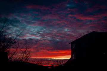 Fiery red and blue sunset clouds roll across the sky above a rural house silhouette. Vivid twilight color, moody atmosphere and layered cloud textures create a dramatic evening backdrop.