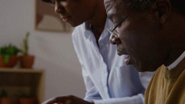 Side view close up of Black senior man carefully pressing buttons on laptop keyboard while working with bills and home finances, young woman helping, copy space