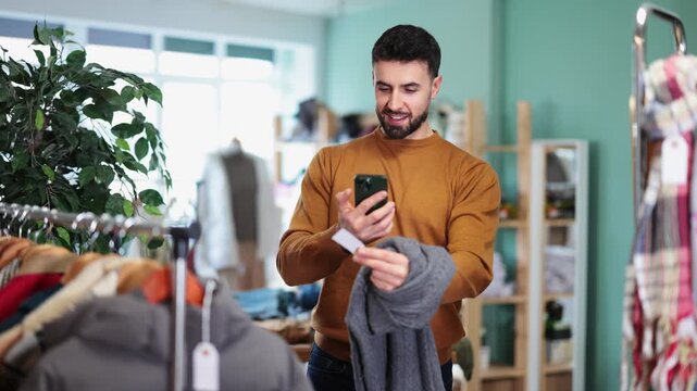  Man scans QR code and label of sweater using mobile phone camera. Customer client is interested in country of origin of product, checks certificate on consumer protection website.