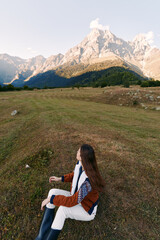 Obraz premium woman sitting on meadow facing mountain landscape in nature, autumn travel scene with long hair, cozy jacket and boots, peaceful outdoor relaxation, panoramic alpine valley and hiking vibe