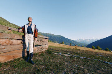 Obraz premium Woman hiking in nature near a wooden fence on a mountain meadow, standing with backpack and boots while enjoying a scenic view and fresh air during a peaceful outdoor adventure.