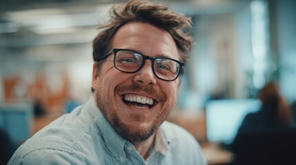 Obraz premium A cheerful man wearing glasses is sitting at his desk in an office environment. He is smiling and appears to be enjoying his work among colleagues in the background.
