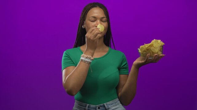 Woman holding a glass bowl of potato chips, hand reaching to grab a chip in studio with purple backdrop and green shirt; snacking pleasure everyday.