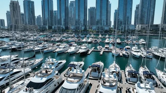 Sweeping aerial drone shot of a massive luxury marina filled with various white sailing boats and speedboats anchored against a dense metropolitan backdrop wealth, harbor, cityscape