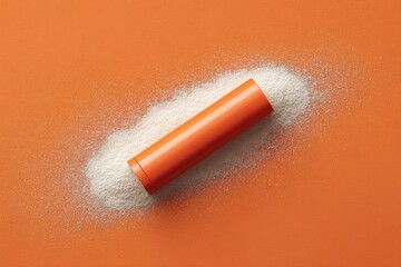 A cylindrical container rests on a pile of granular material against an orange background