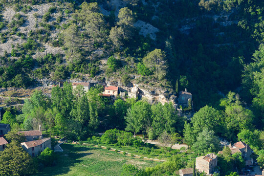 Aerial view of rustic stone houses with red roofs set against a forested limestone hillside in Navacelles. Bright sunlight brings out the vibrant green foliage and rugged textures of the landscape.