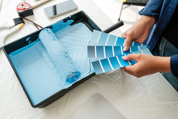 Fototapeta na wymiar A person holds a fan of paint swatches next to a blue roller in a tray, preparing for a home makeover. The image conveys planning, creativity, and color selection in a stylish interior project.