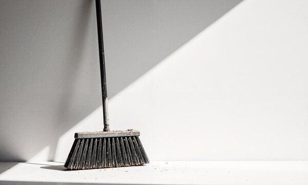 Sweeping Tool Stillness. A wide, industrial push broom leans against a flawless white cyc wall, its bristles frayed and dusted