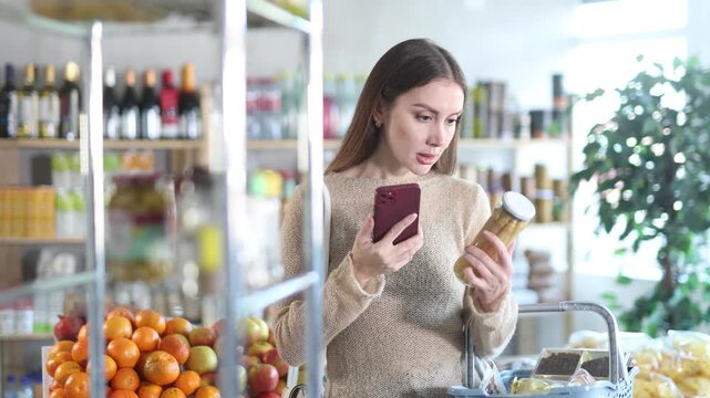 Interested young woman making purchases in local grocery store, using convenient mobile app to pay online for pickled asparagus by scanning barcode on jar