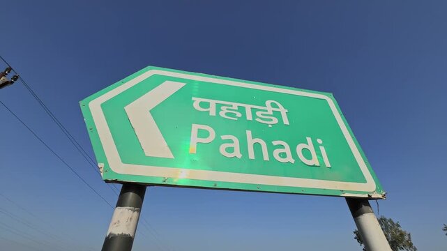 Orbiting view of a green highway direction sign reading Pahadi in Hindi and English, mounted on metal poles against a clear blue sky, showing rural Indian road signage.