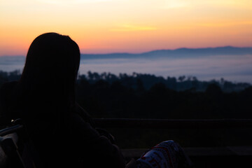 Naklejka premium Back view silhouette of woman sitting and watching sea of mist over mountain valley at sunrise, peaceful morning landscape from wooden balcony, concept of freedom, soul searching and slow travel.